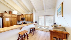 Una cocina con armarios de madera, microondas y un ventanal con persianas. Hay dos taburetes de madera y una pequeña mesa con papeles.