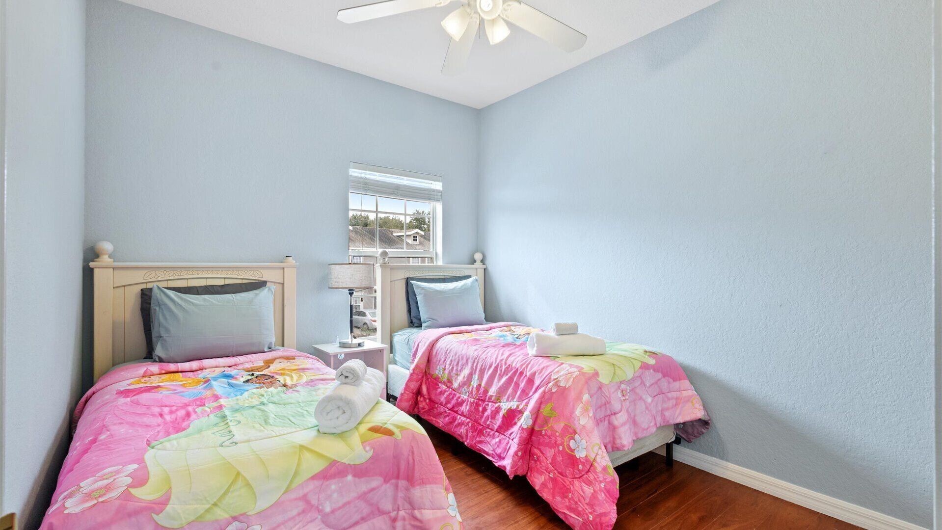 A bedroom with two beds, each with a floral bedspread, a nightstand, a lamp, and a window with a view of houses.