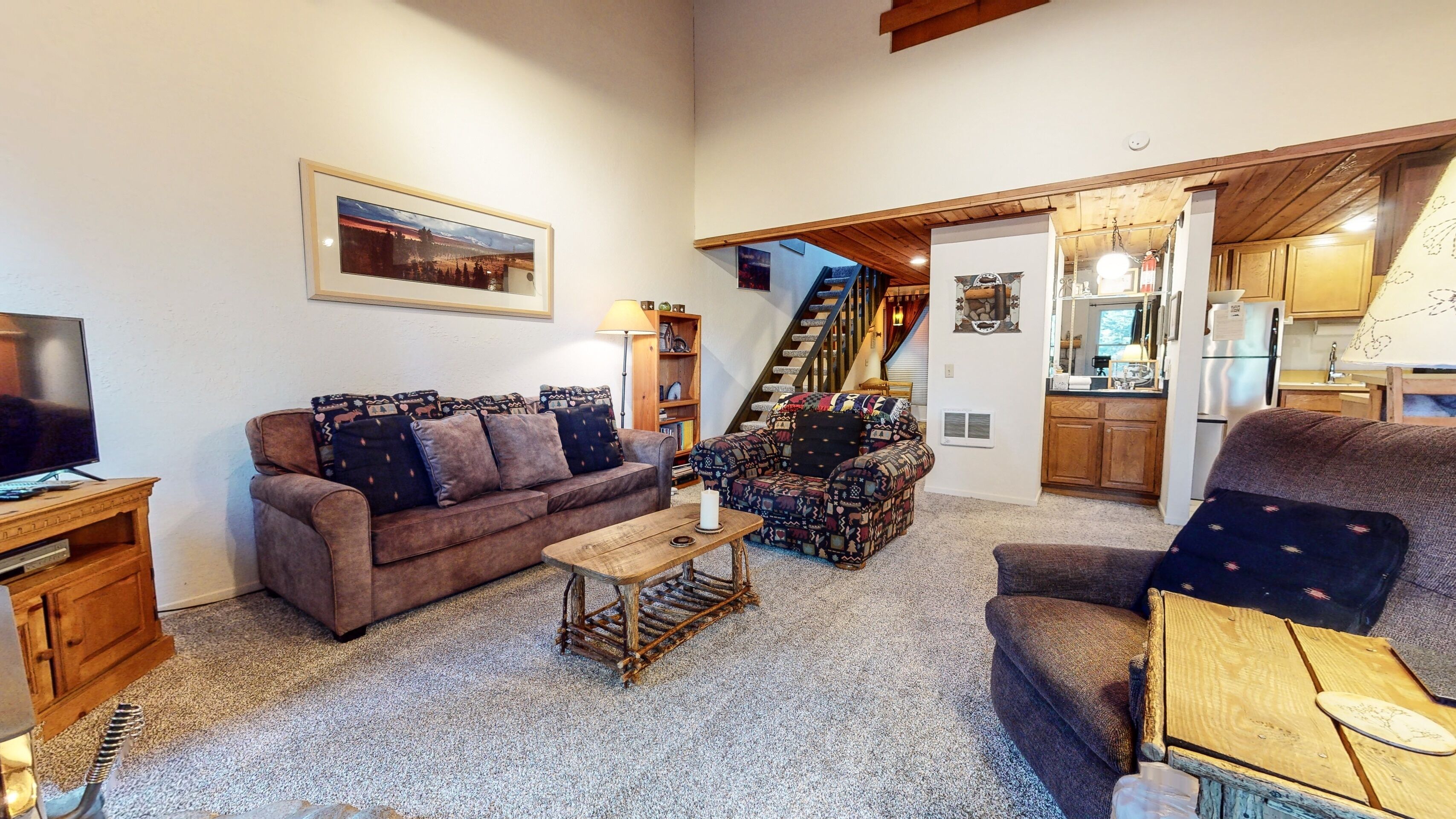 A cozy living room with a brown sofa, patterned armchairs, a coffee table, and a television. A staircase leads to a kitchen with wooden cabinets and a refrigerator.