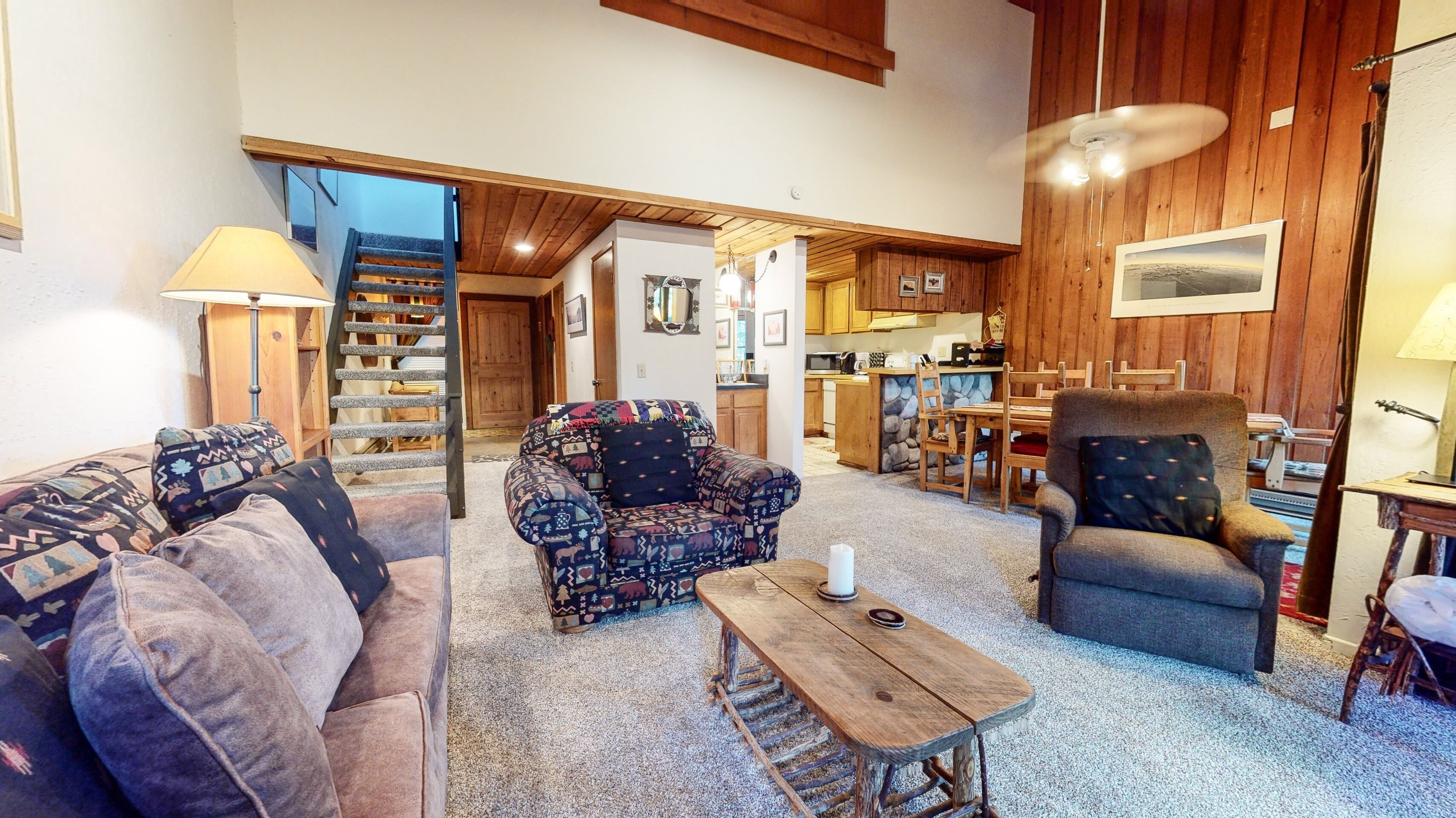 A cozy living room with a sofa, armchair, and coffee table, a staircase leading to a kitchen, and wooden walls.