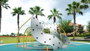 A playground with climbing structures and a slide, surrounded by palm trees and a building in the background. - Solana #1 - 7 Bed Villa w/ Private Pool (Davenport)