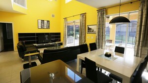 A modern living room with a yellow wall, a black sofa, a dining table with chairs, a TV, and a large window with curtains. - Solana #1 - 7 Bed Villa w/ Private Pool (Davenport)