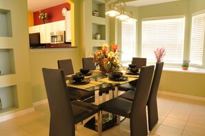 A dining area with a glass table and black chairs, a vase of flowers, and a kitchen in the background.