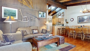A cozy living room with a sofa, coffee table, and wooden chairs. There are framed pictures on the wall, a lamp, and a chandelier.