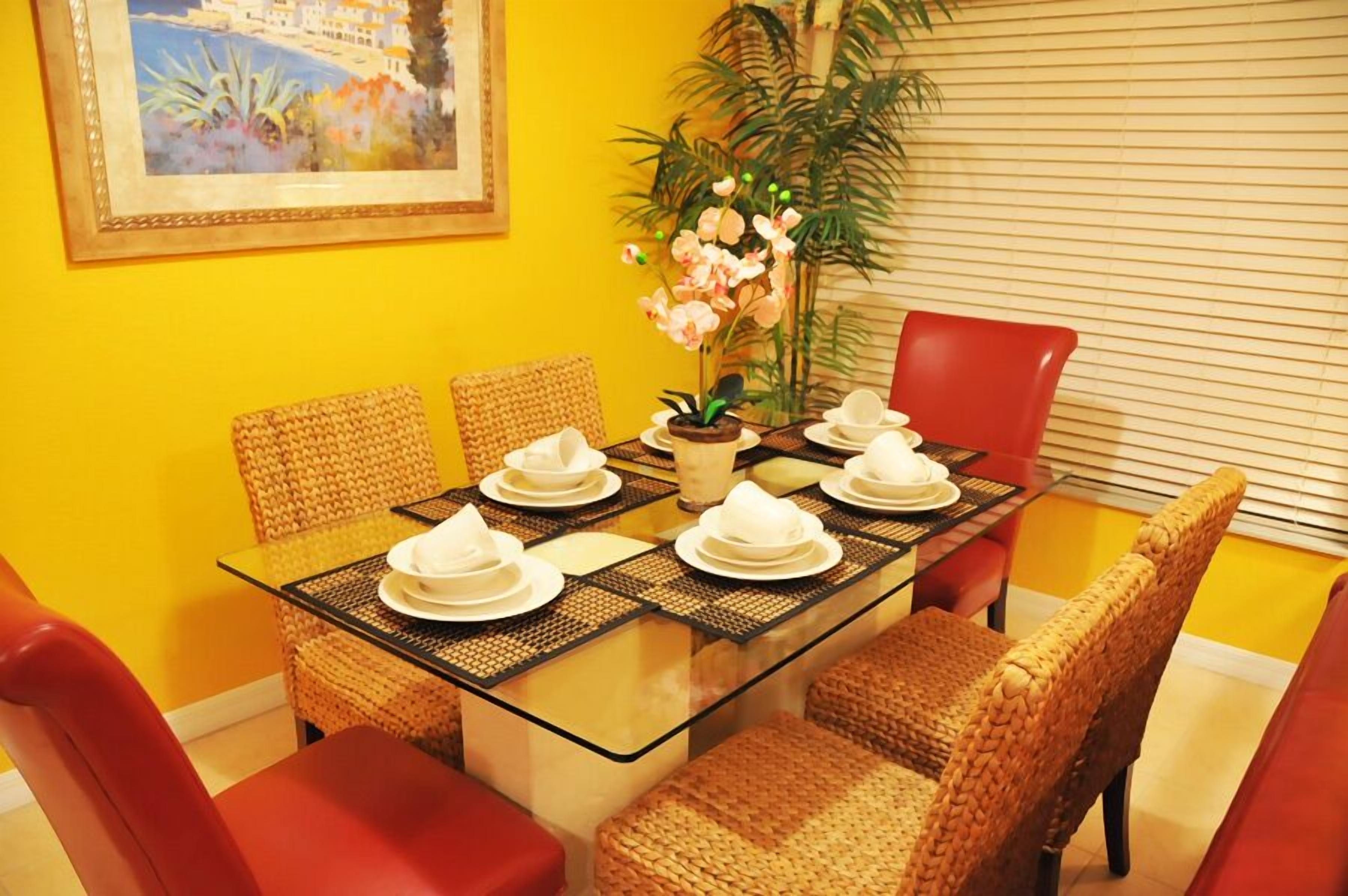 A dining area with a glass table, wicker chairs, and a red chair. There is a plant and a framed picture on the wall.
