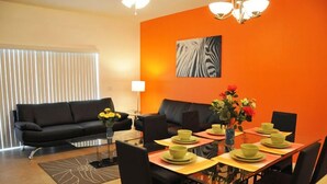 Ein modernes Wohnzimmer mit einem schwarzen Sofa, einem Esstisch mit grünen Tellern und einem Zebrastreifen-Kunstwerk an der orangen Wand.