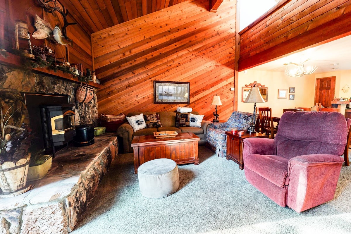 A cozy cabin interior with a stone fireplace, wooden walls, a dining area, and a kitchen.