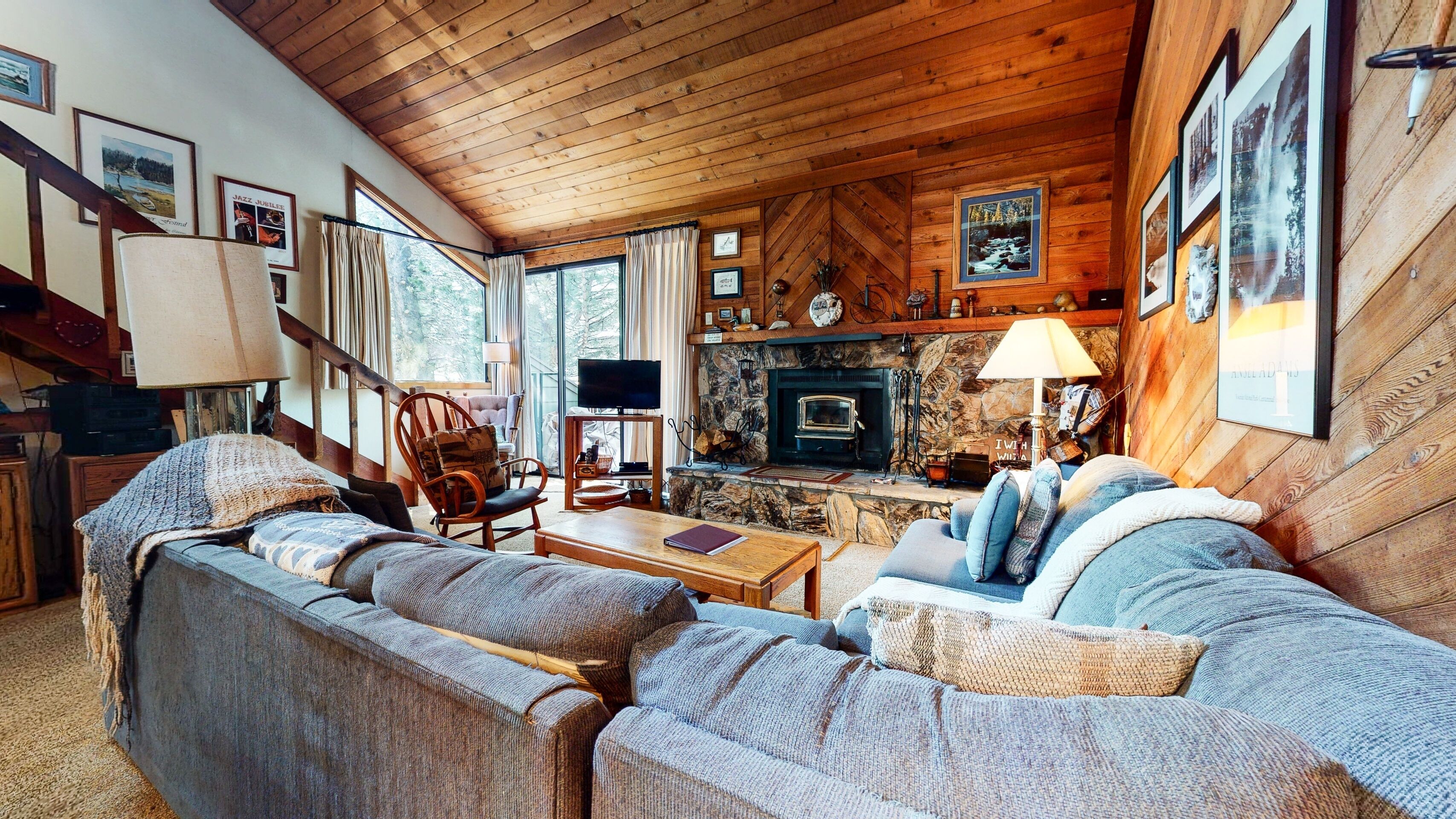 A cozy cabin interior with a stone fireplace, wooden furniture, and a staircase leading to an upper level.
