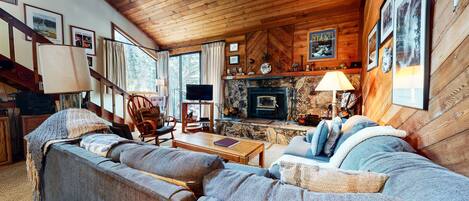 A cozy cabin interior with a stone fireplace, wooden furniture, and a staircase leading to an upper level.