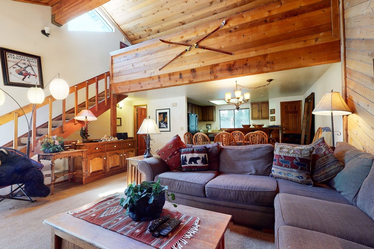 A cozy living room with a wooden ceiling, a staircase, a dining area, and a kitchen in the background.