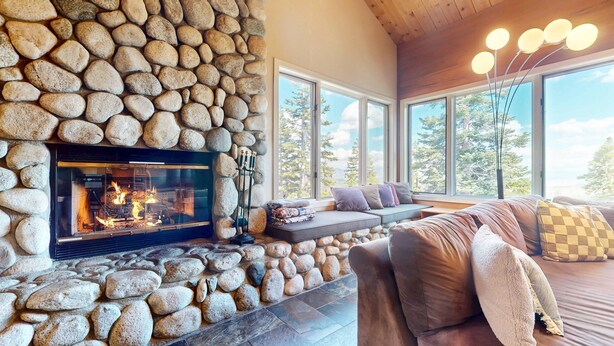 A stone fireplace with a fire, a stone wall, a sofa with cushions, and large windows with a view of trees. - Bridges Townhome 13 Mountain View Townhome just Steps to the Slopes with Private Garage and Washer Dryer by RedAwning (Mammoth Lakes)