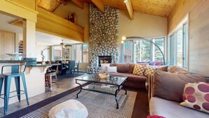 A cozy living room with a stone fireplace, a dining area with a high wooden ceiling, and large windows offering a view of trees. - Bridges Townhome 13 Mountain View Townhome just Steps to the Slopes with Private Garage and Washer Dryer by RedAwning (Mammoth Lakes)