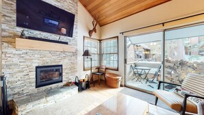 Un salon chaleureux avec une cheminée en pierre, un grand téléviseur fixé au mur et une vue sur l’extérieur grâce à de grandes fenêtres.
