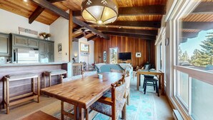 A rustic cabin interior with a wooden dining table, benches, a kitchen area with a refrigerator, and a large window offering a view of trees.