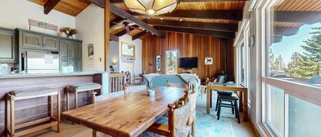 A rustic cabin interior with a wooden dining table, benches, a kitchen area with a refrigerator, and a large window offering a view of trees.