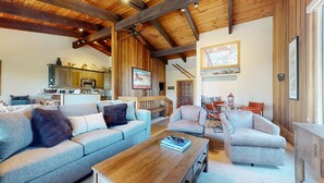A spacious living area with a high ceiling, wooden beams, and a cozy fireplace. There is a kitchen in the background with modern appliances and a dining area with a table and chairs.