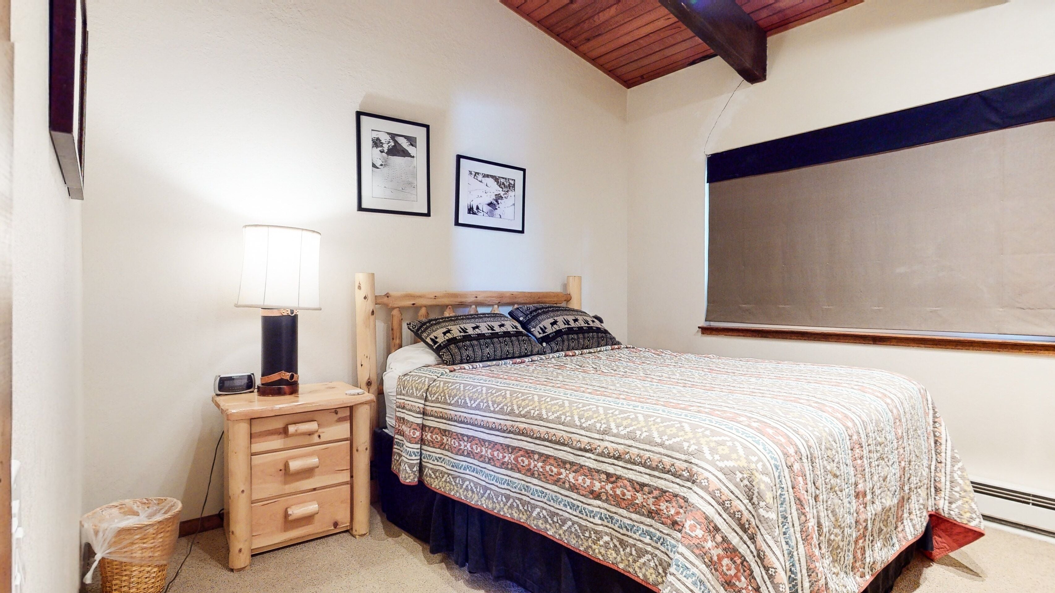 Ein Schlafzimmer mit einem hölzernen Bett, gemusterten Betttextilien, einer Nachttischlampe, einem kleinen Nachttisch mit Schubladen und zwei gerahmten Bildern an der Wand.
