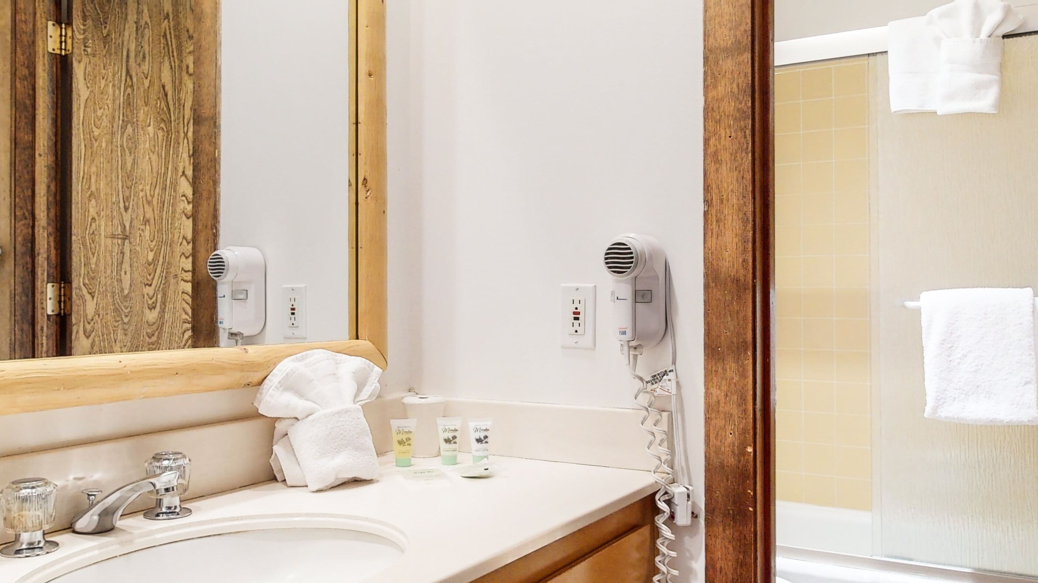 A bathroom with a wooden-framed mirror, a white sink with a towel, and two hair dryers mounted on the wall.