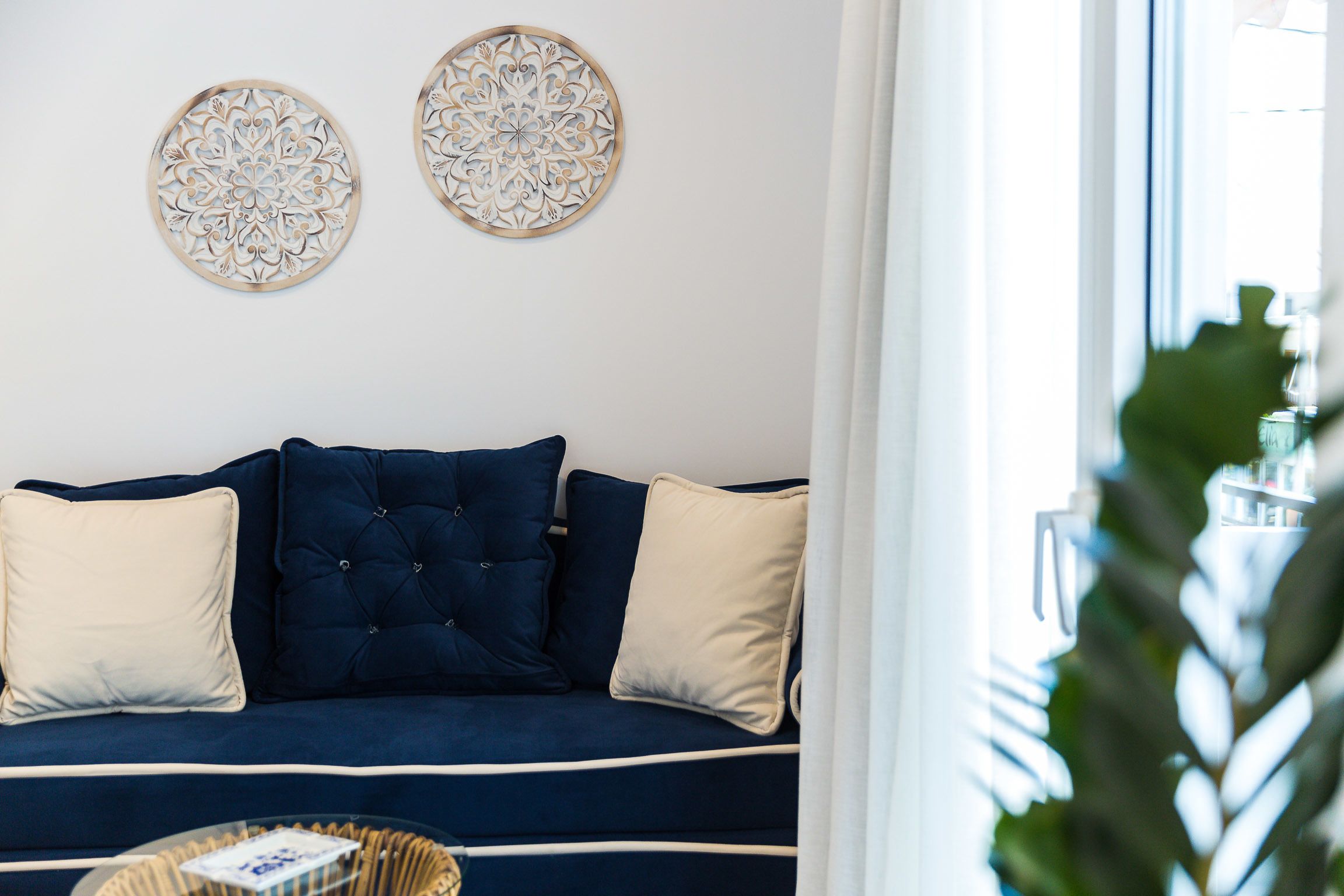 A dark blue sofa with white and beige cushions, two decorative wall pieces, and a wicker side table with a book.