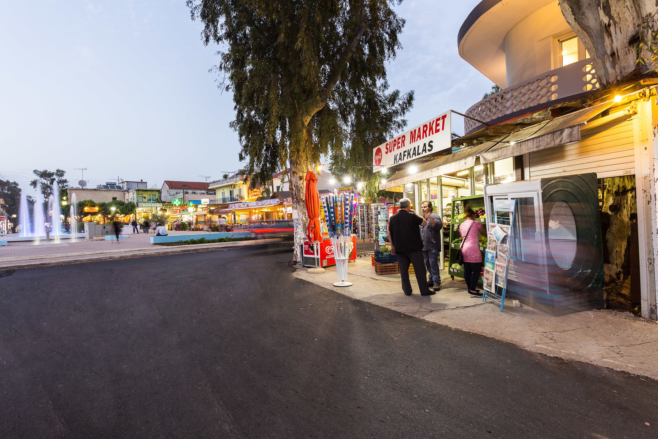 Straßenszene mit einem Supermarkt-Schild, einer Kiosk und einem Brunnen.
