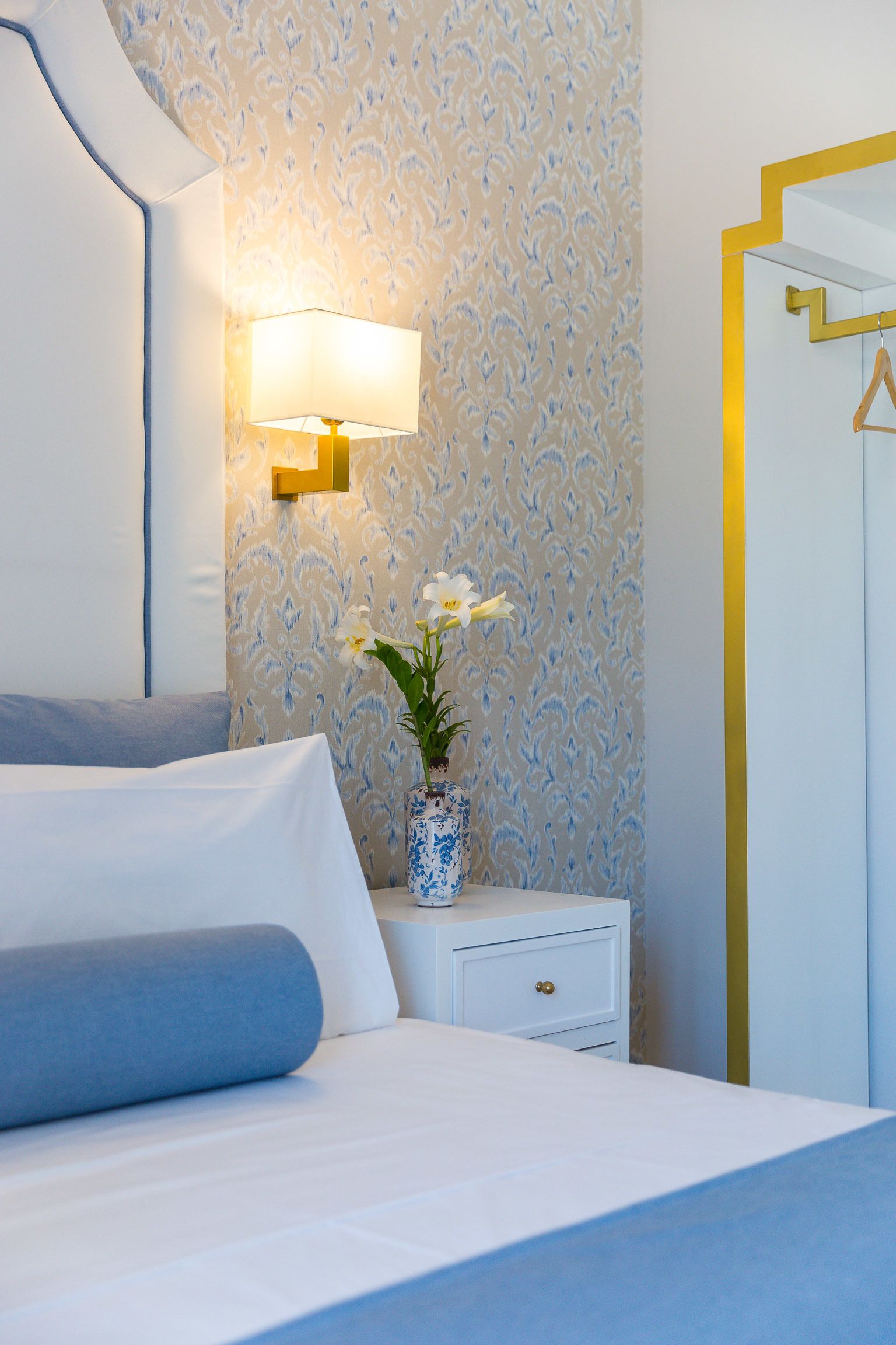 Ein ordentlich bezogenes Bett mit blauem Kopfkissen, ein Nachttisch mit Blumen in einer Vase und eine Wand mit Tapete mit Muster sowie einem goldfarbenen Spiegel.