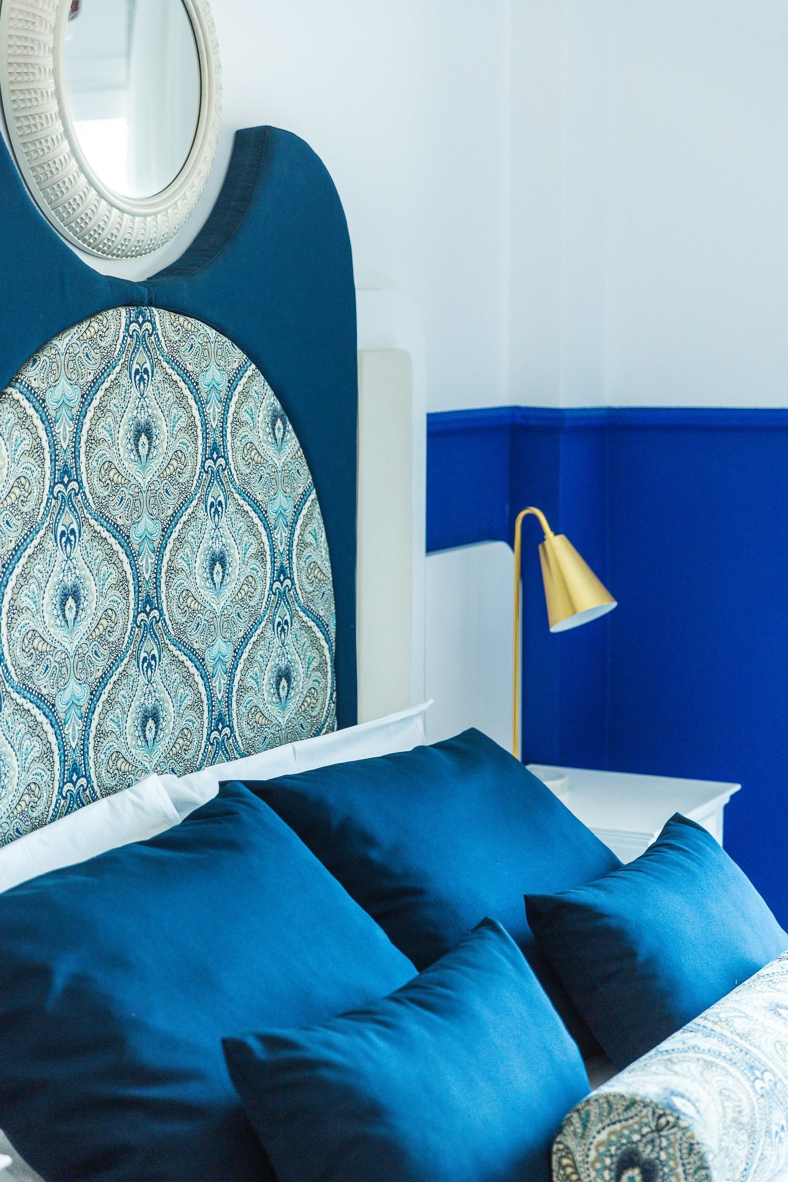 A bed with blue bedding, a round mirror, and a bedside lamp.