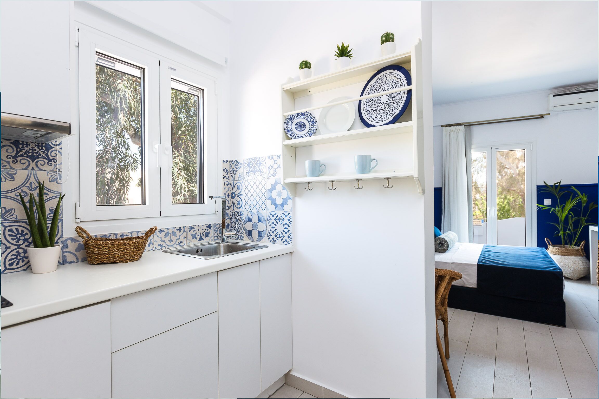 Una cocina moderna con armarios blancos, un lavamanos y una ventana. Se ven repisas con platos y tazas, además de una cesta de mimbre sobre la encimera.