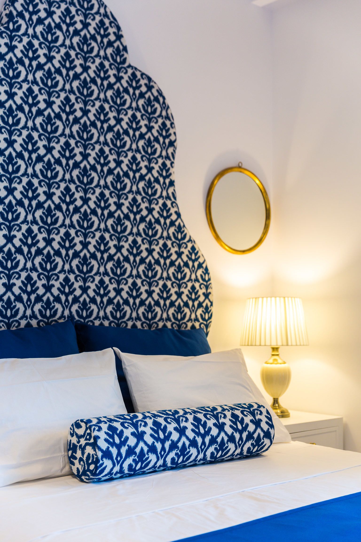 Een bed met een hoofdbord met patroon, een bedlampje en een spiegel aan de muur.