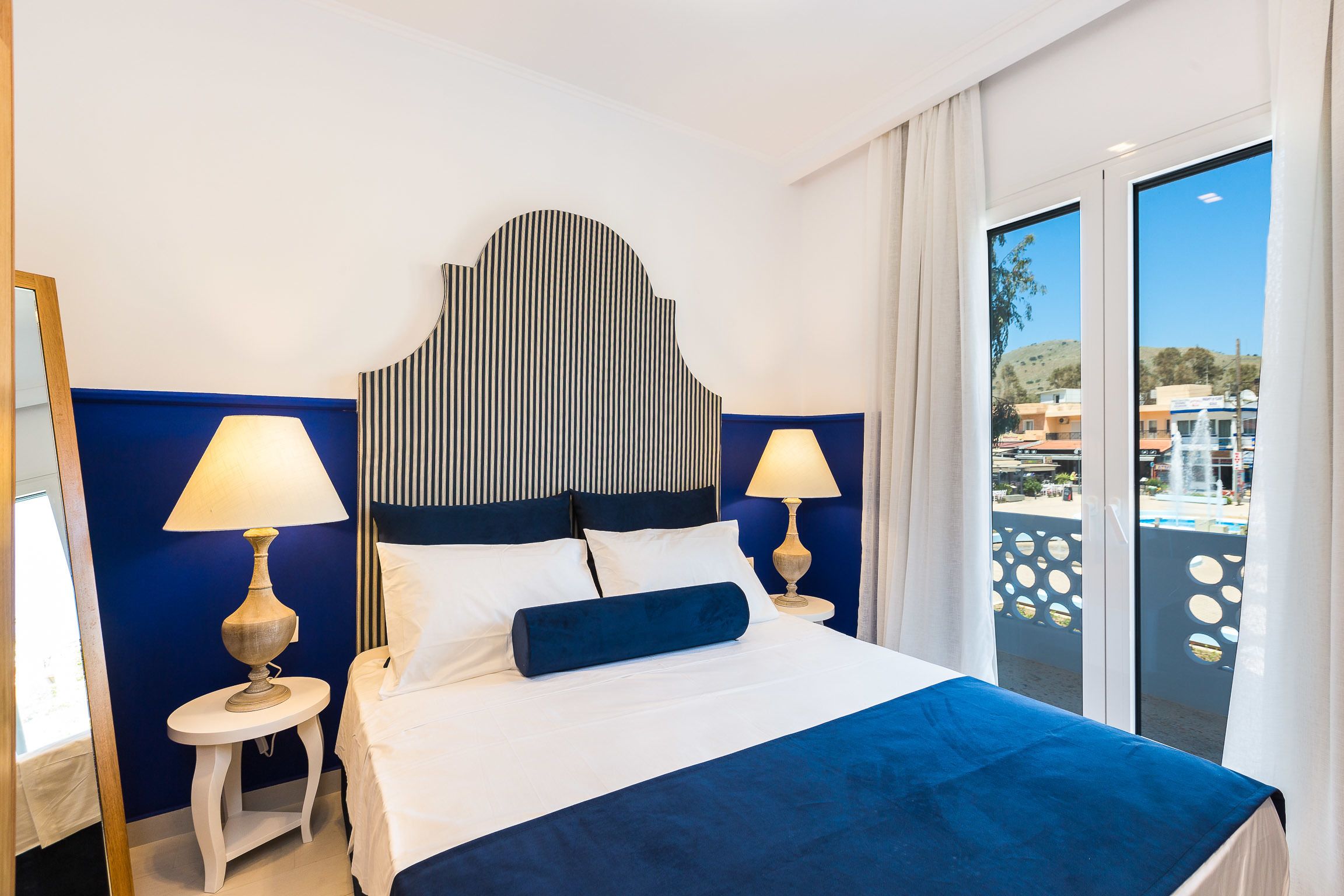 A hotel room with a bed, two bedside tables with lamps, a striped headboard, and a view of buildings through a large window.