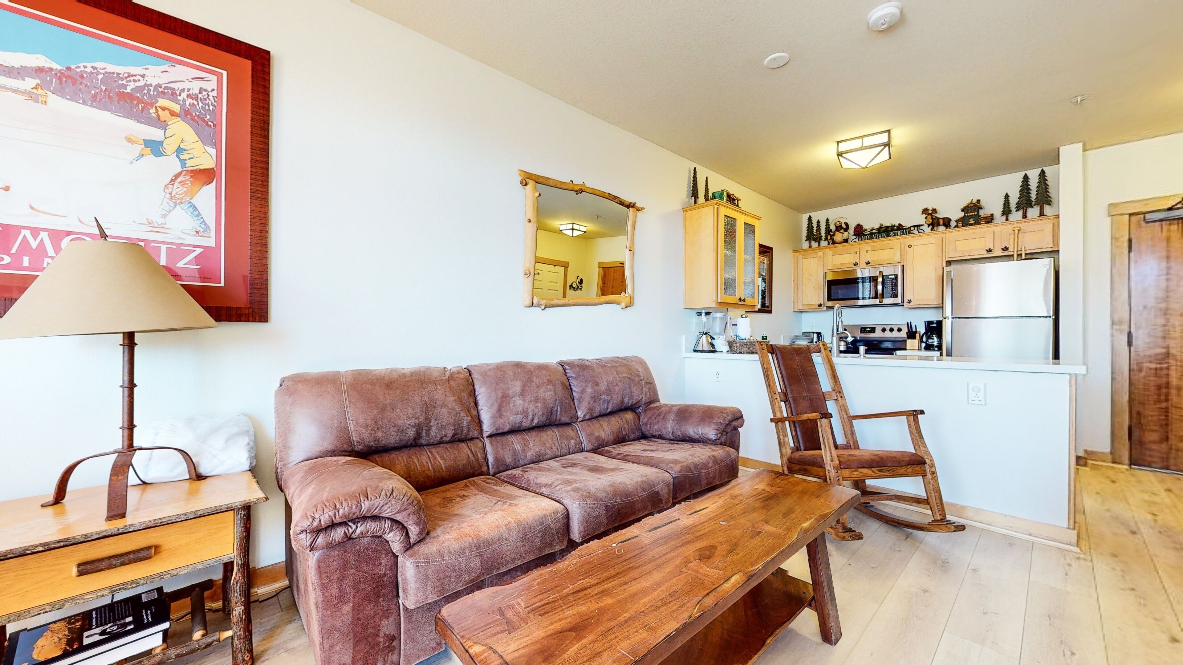 A cozy living room with a brown leather sofa, a wooden bench, and a rustic lamp. A kitchen with modern appliances and a wooden island is visible in the background.