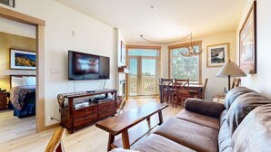 A living room with a flat-screen TV, wooden furniture, a dining area with a table and chairs, and a balcony with a view. - Sunstone 103 Updated Condo At Sunstone Lodge with Great Complex Amenities, near Ski-In Ski-Out by RedAwning (Mammoth Lakes)