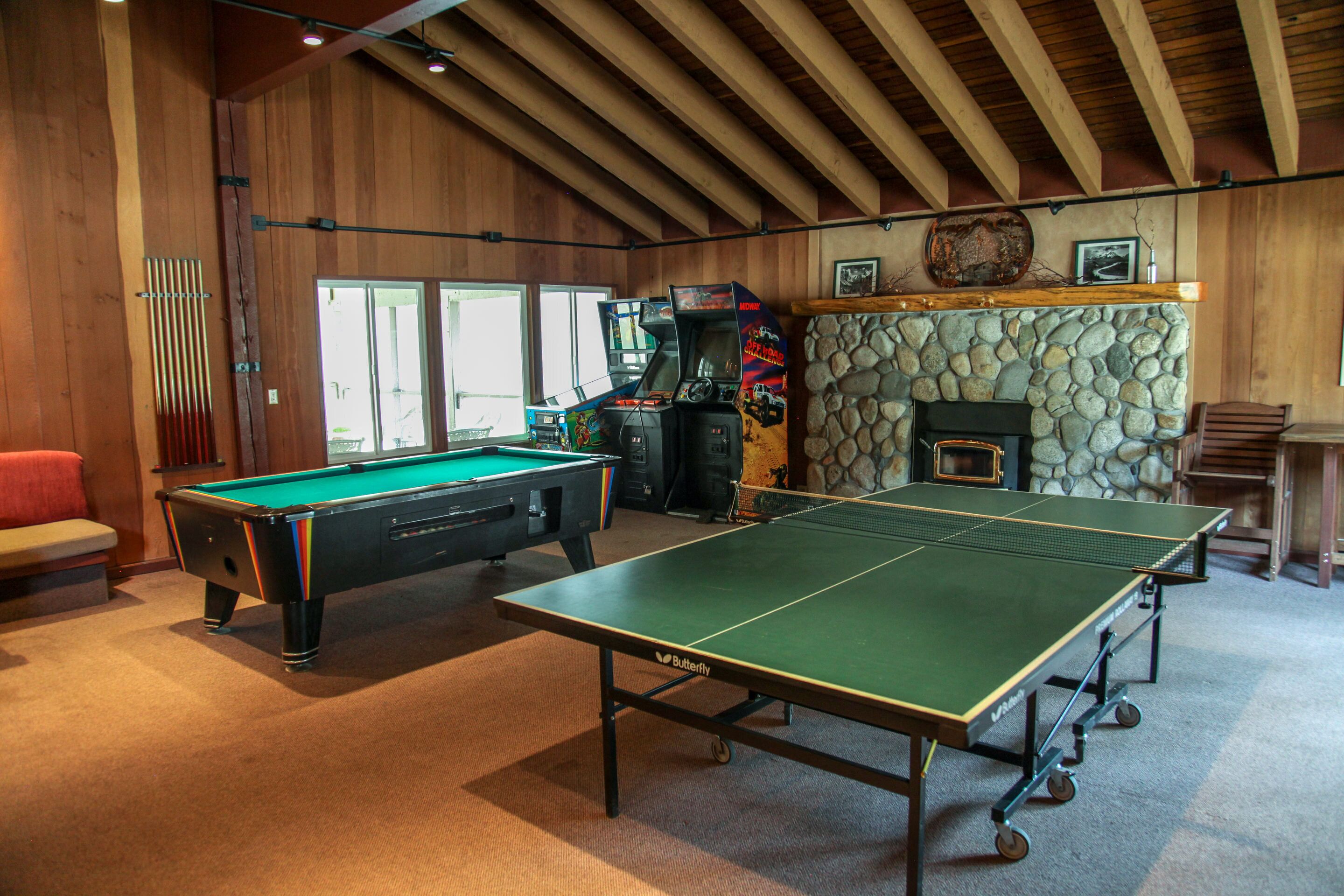 A pool table, a ping pong table, and a billiards table in a room with wooden walls and a stone fireplace.