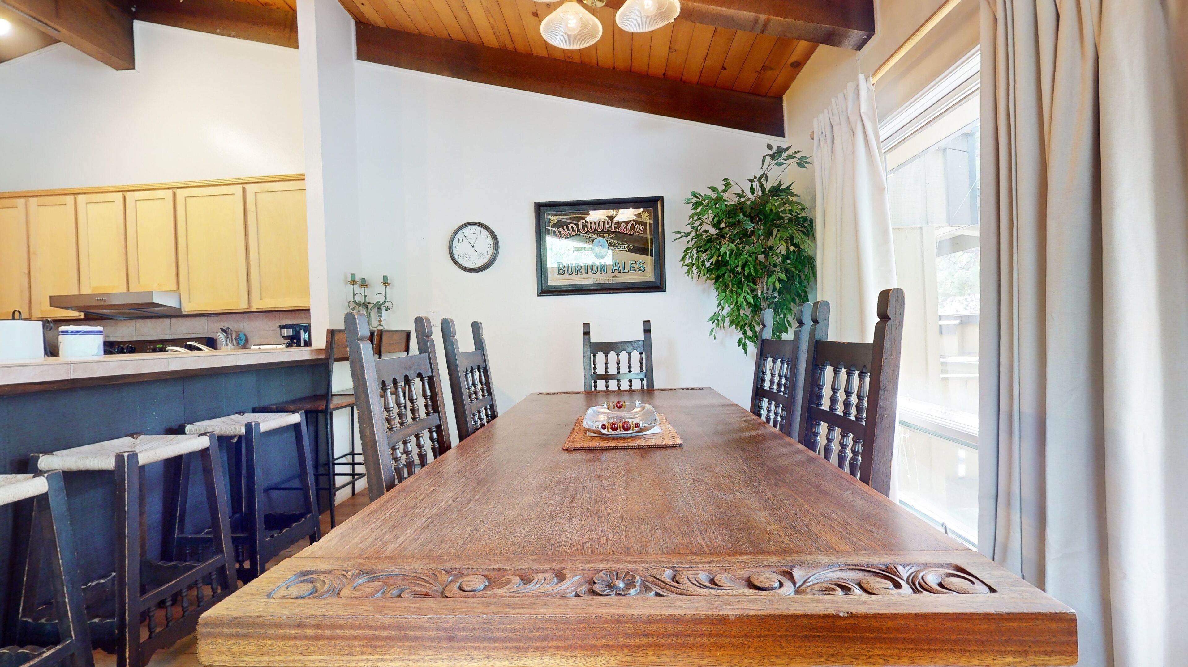 A wooden dining table with ornate carvings, surrounded by chairs, in a kitchen with wooden cabinets and a hanging plant.