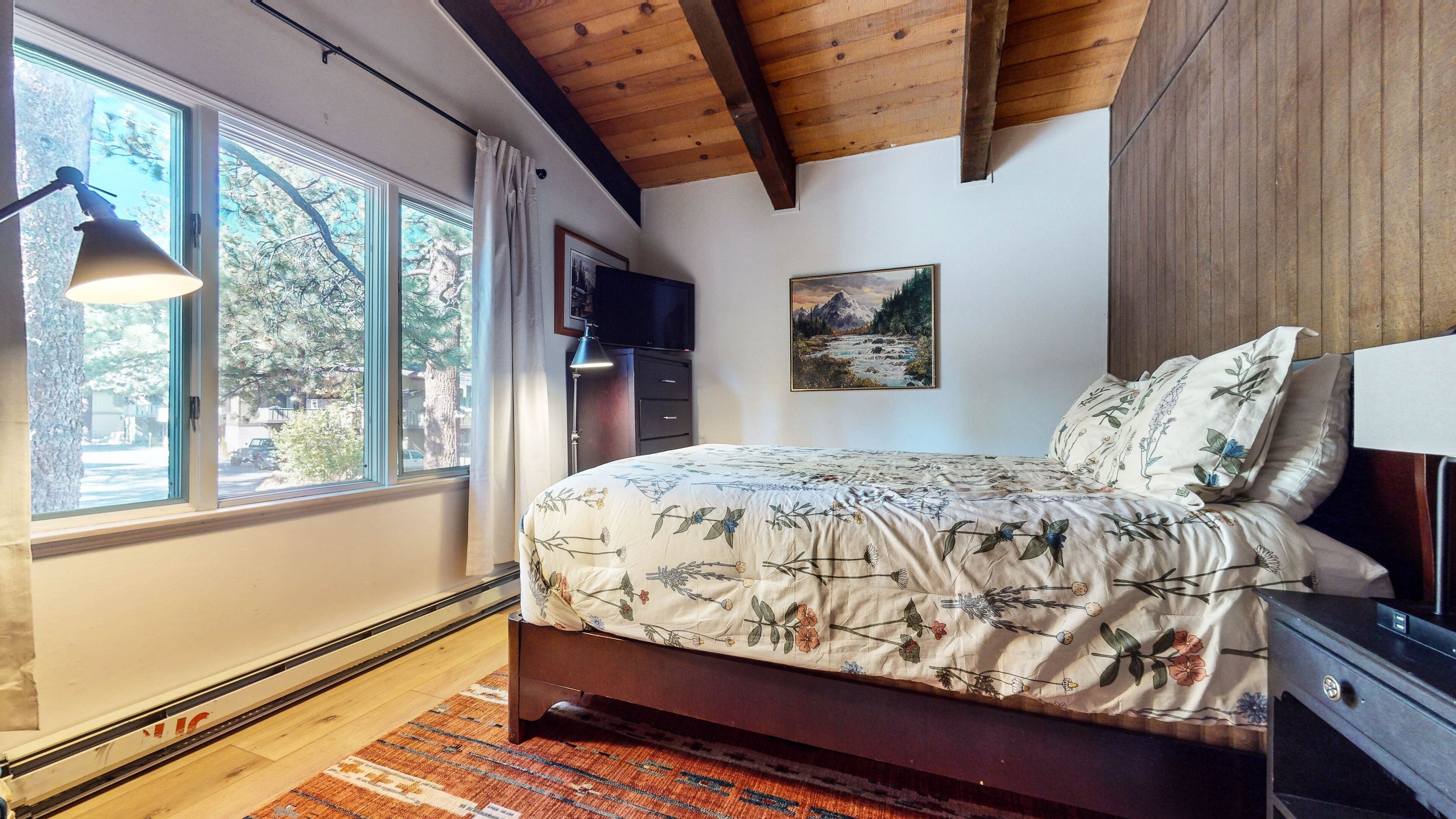 Un dormitorio con techo de madera, cama con ropa de cama floral, televisor sobre un mueble bajo y una ventana con cortinas.