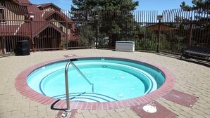 Un bain à remous entouré d’une terrasse en briques, délimité par une clôture en métal, avec un bâtiment visible au loin.