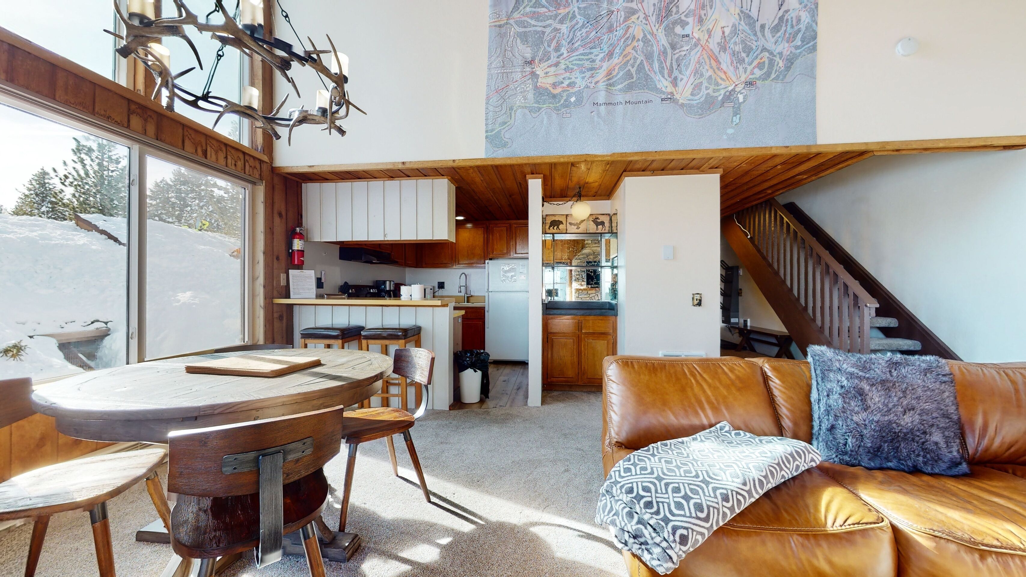 A modern kitchen with wooden cabinets, a dining area with a round table and chairs, a leather sofa with patterned cushions, and a staircase leading to an upper level.