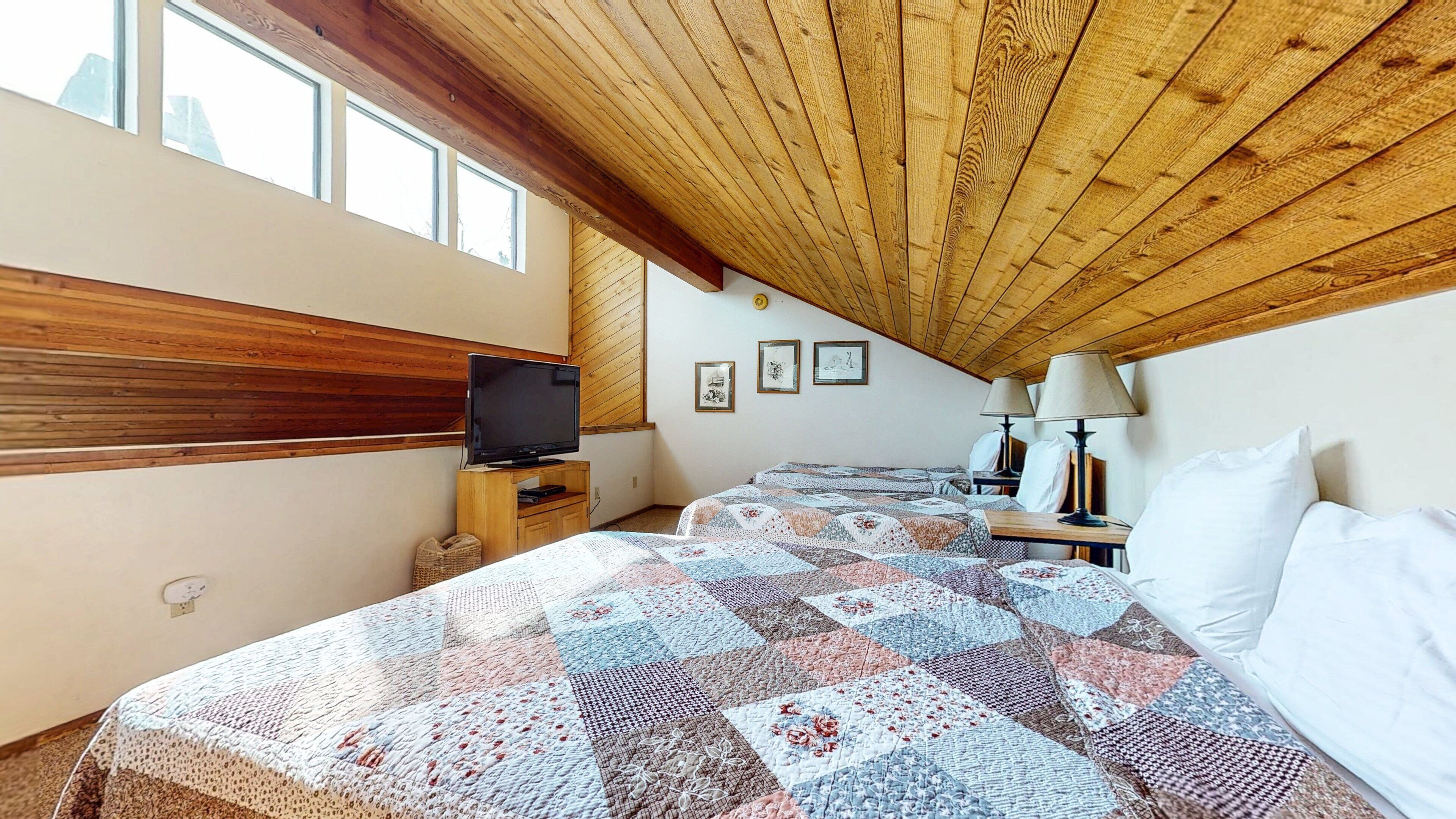 A cozy bedroom with a wooden ceiling, a television, two beds with quilts, bedside lamps, and framed pictures on the wall.