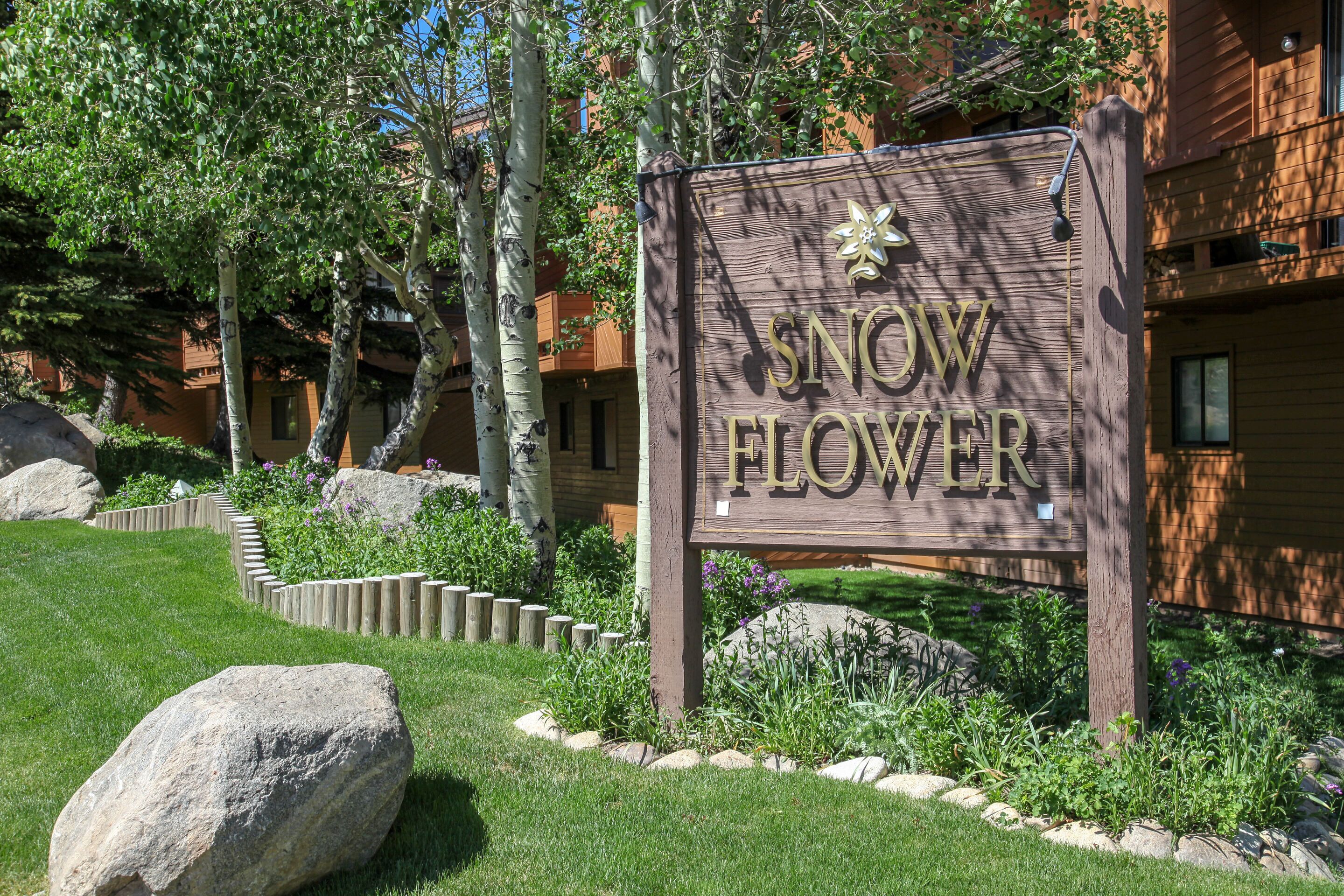 Ein hölzernes Schild mit der Aufschrift 'SNOW FLOWER' in goldener Schrift, umgeben von einem angelegten Garten mit großen Steinen und Bäumen.