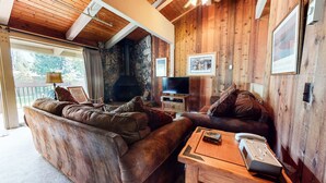 Interior acolhedor de uma cabana com lareira de pedra, paredes de madeira, um sofá grande e uma televisão.