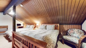 Quarto com teto de madeira, uma cama grande com roupa de cama estampada, uma poltrona com tecido floral e uma mesa de cabeceira com abajur.