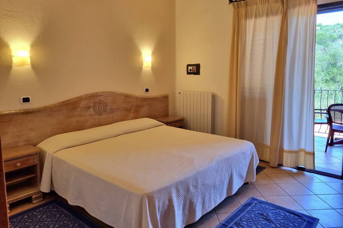 Une chambre d’hôtel avec un lit, une table de chevet et une vue sur l’extérieur par une fenêtre munie de rideaux.