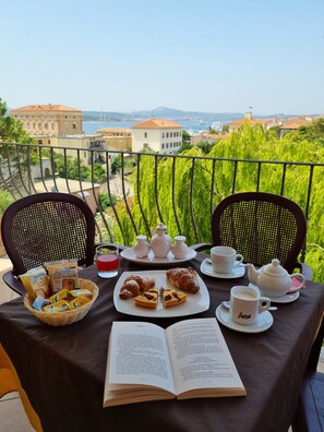 Een balkon met een tafel, belegd met een boek, gebak en een theepot, met uitzicht op een stadsgezicht.