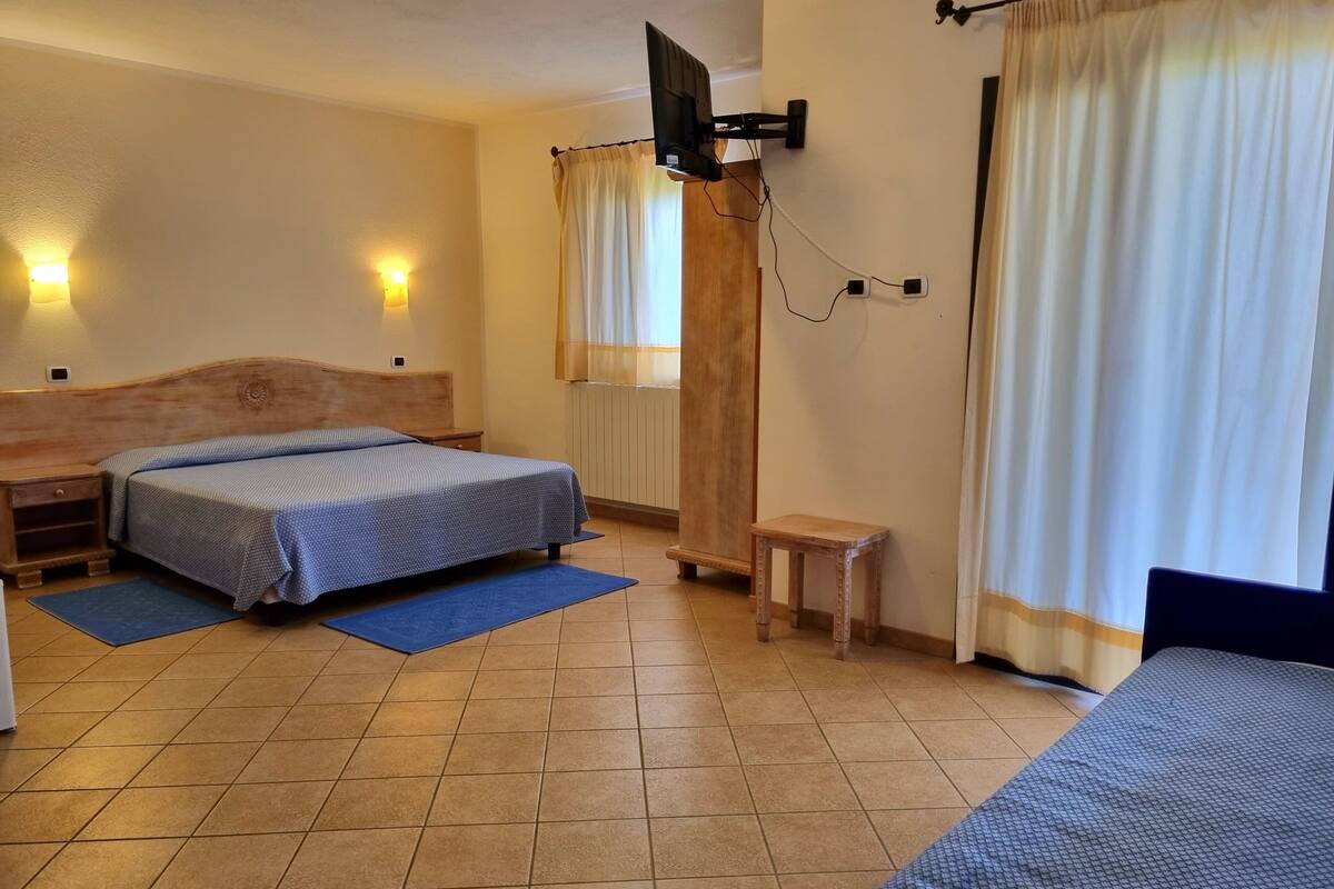 Une chambre d’hôtel avec deux lits, un téléviseur fixé au mur, un petit tabouret en bois et une fenêtre avec des rideaux.