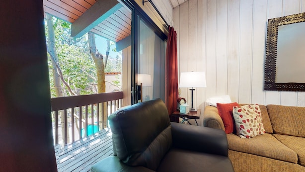 Una habitación acogedora con techo de madera, un sillón mullido, un sofá con cojines estampados, una mesita de noche con lámpara y un espejo grande en la pared.