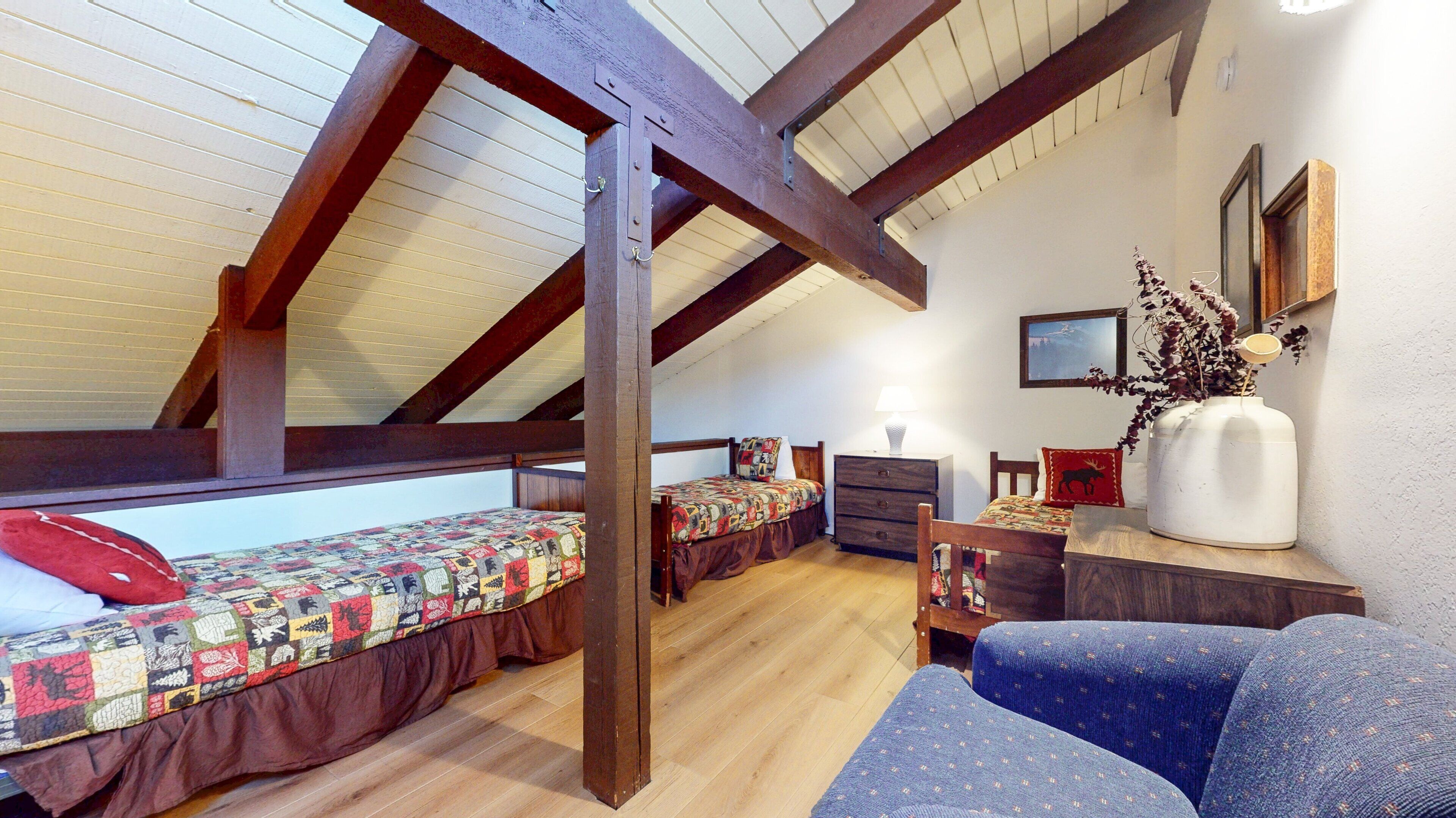 A cozy attic bedroom with wooden beams, a quilted bed, a dresser, and a chair.