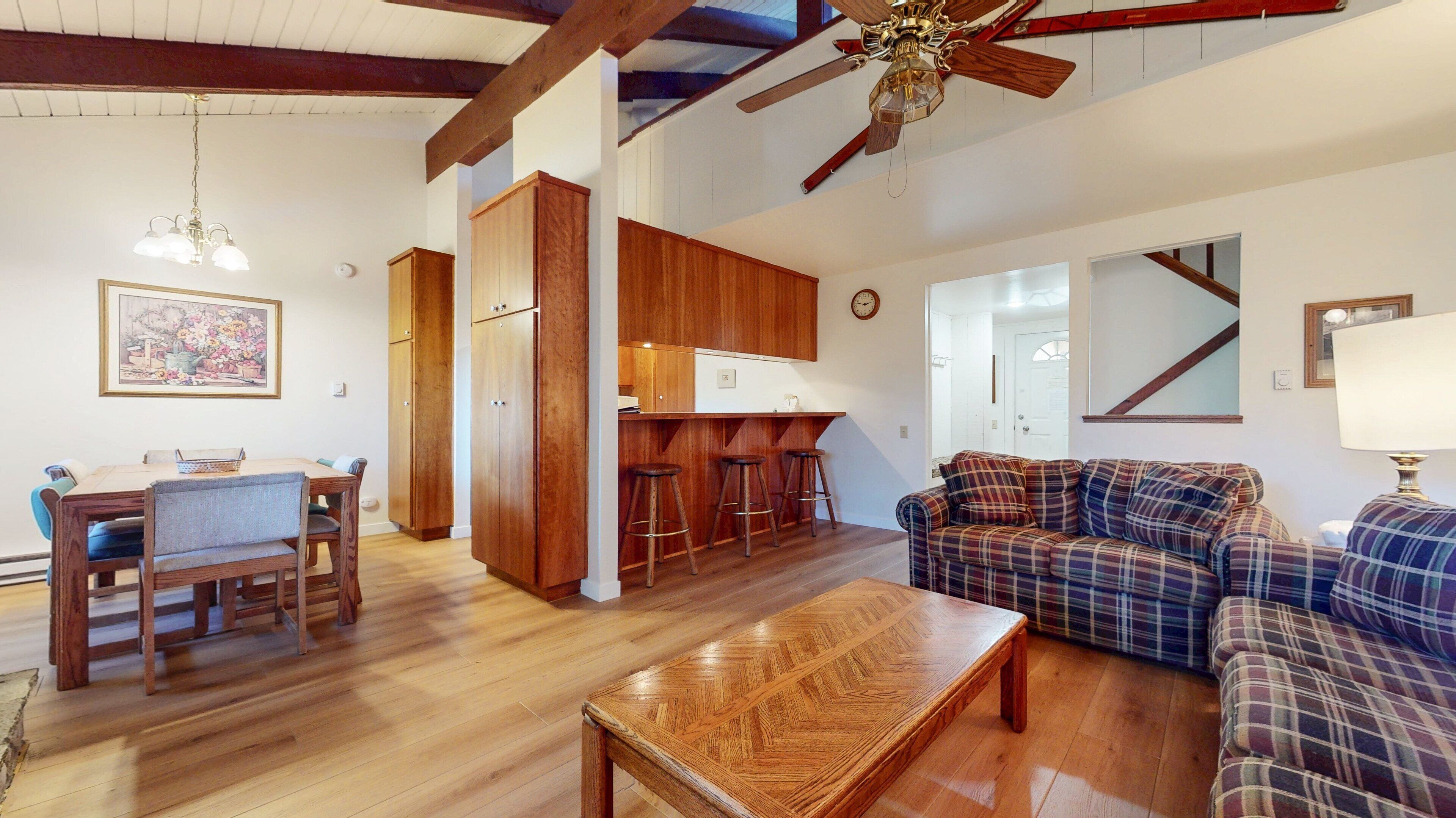 A spacious living area with a wooden coffee table, a plaid sofa, a dining table with chairs, and a kitchen area with stools.