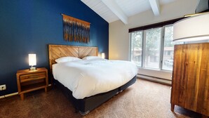 Un dormitorio con una cama grande, una mesita de noche con lámpara, cabecero de madera y ventana con vistas al exterior.