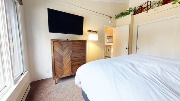Un dormitorio con una cama, un televisor montado en la pared, un armario de madera y una puerta que da al baño.