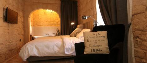 A bedroom with a stone wall, a bed, a chair with a cushion, and a mirror.