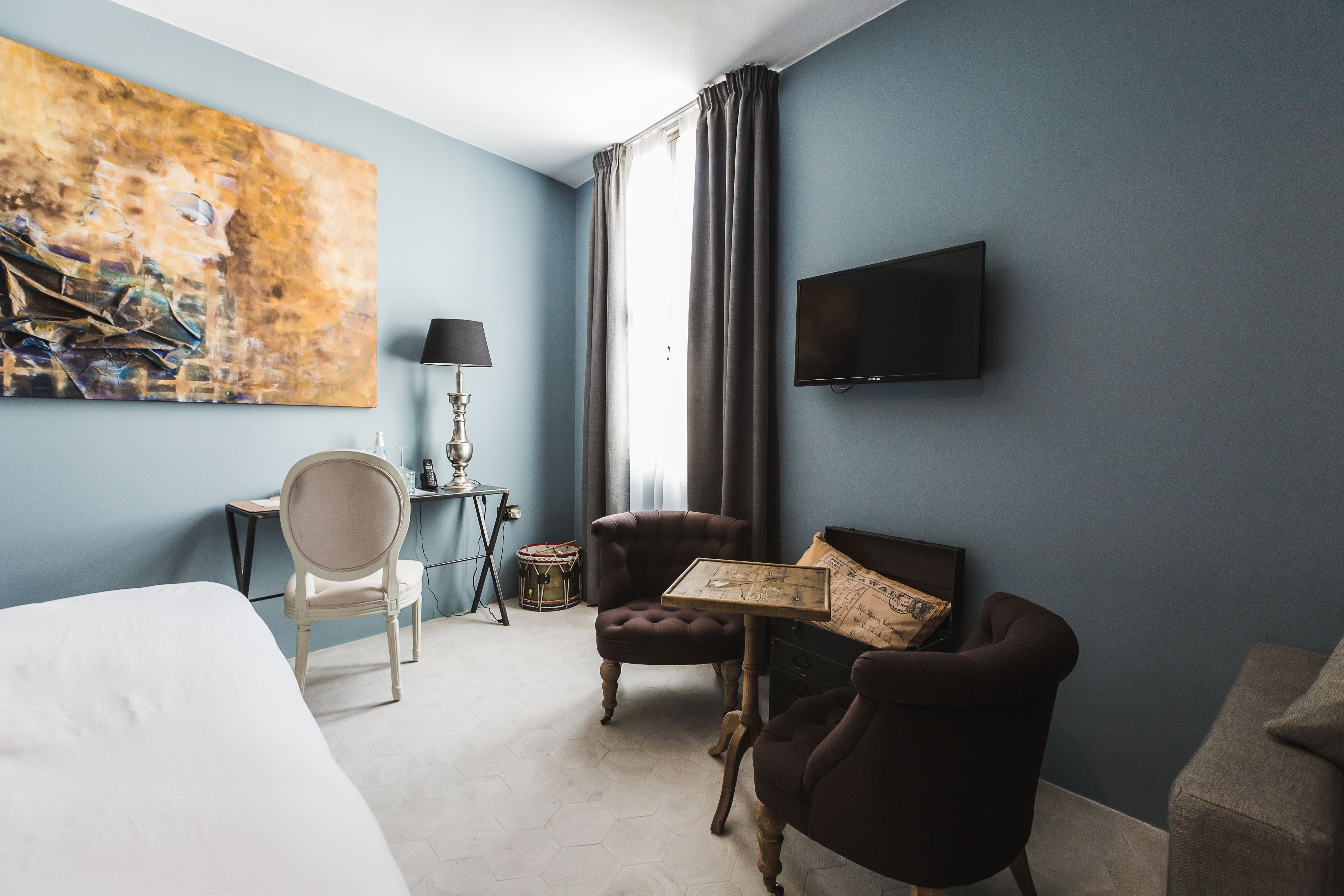 Ein Hotelzimmer mit einem Bett, zwei Sesseln, einem kleinen Tisch, einer Lampe, einem Fernseher und einem großen abstrakten Gemälde an der Wand.